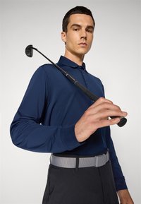Homme en polo à manches longues navy et ceinture grise tient un club de golf reposant sur son épaule contre un fond uni.