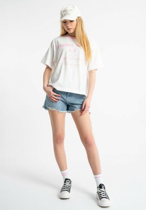 Młoda kobieta w białym czapce, oversize białej koszulce z różowym napisem, podartych niebieskich jeansowych szortach, białych skarpetkach i czarnych trampkach stoi na białym tle.