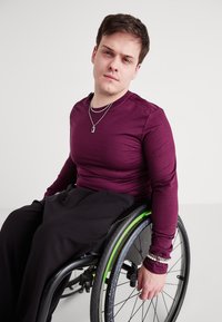 Mélykék, hosszú ujjú, testre simító ing fekete nadrággal párosítva. Kerekesszékben ül, zöld részletekkel ellátott kerékkel, réteges nyakláncokat visel.