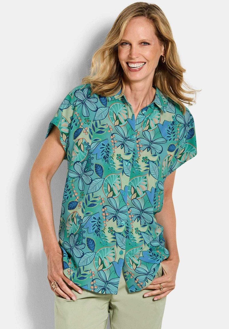 Femme souriante, portant une chemise à manches courtes avec des motifs floraux et de feuilles bleus et verts, ainsi qu'un pantalon beige clair.