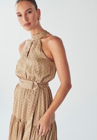 Tan polka dot -mekko, jossa on halterneck-olkaimet ja avonainen leikkaus. Mekossa on vyötäröllä vyö, jossa on pyöreä solki ja kerroksellinen hame.