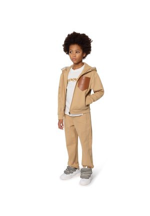 Sweat à zip beige avec un pantalon beige et un t-shirt blanc. Le sweat présente une patch en cuir marron. Des baskets blanches et noires texturées complètent la tenue.