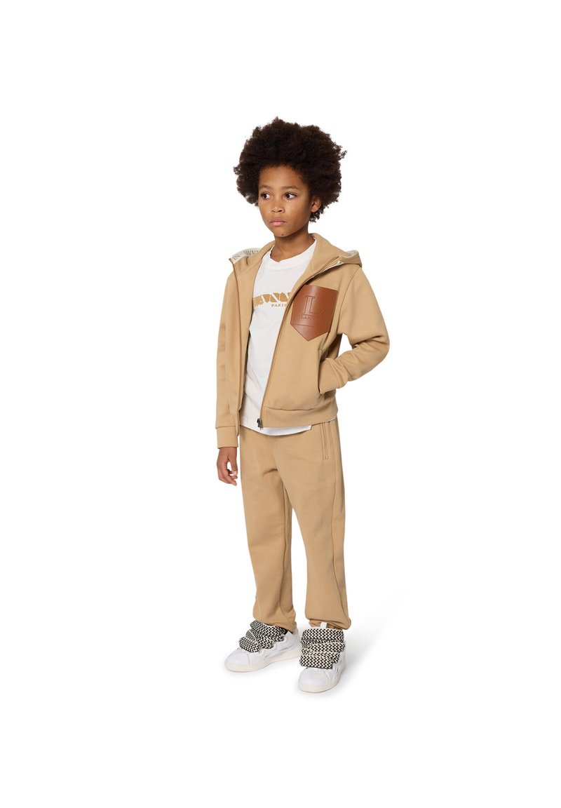 Beige Kapuzenpulli mit hellen Hosen und einem weißen T-Shirt. Der Kapuzenpulli hat ein braunes Leder-Patch. Weiße und schwarze Sneakers mit Textur runden das Outfit ab.