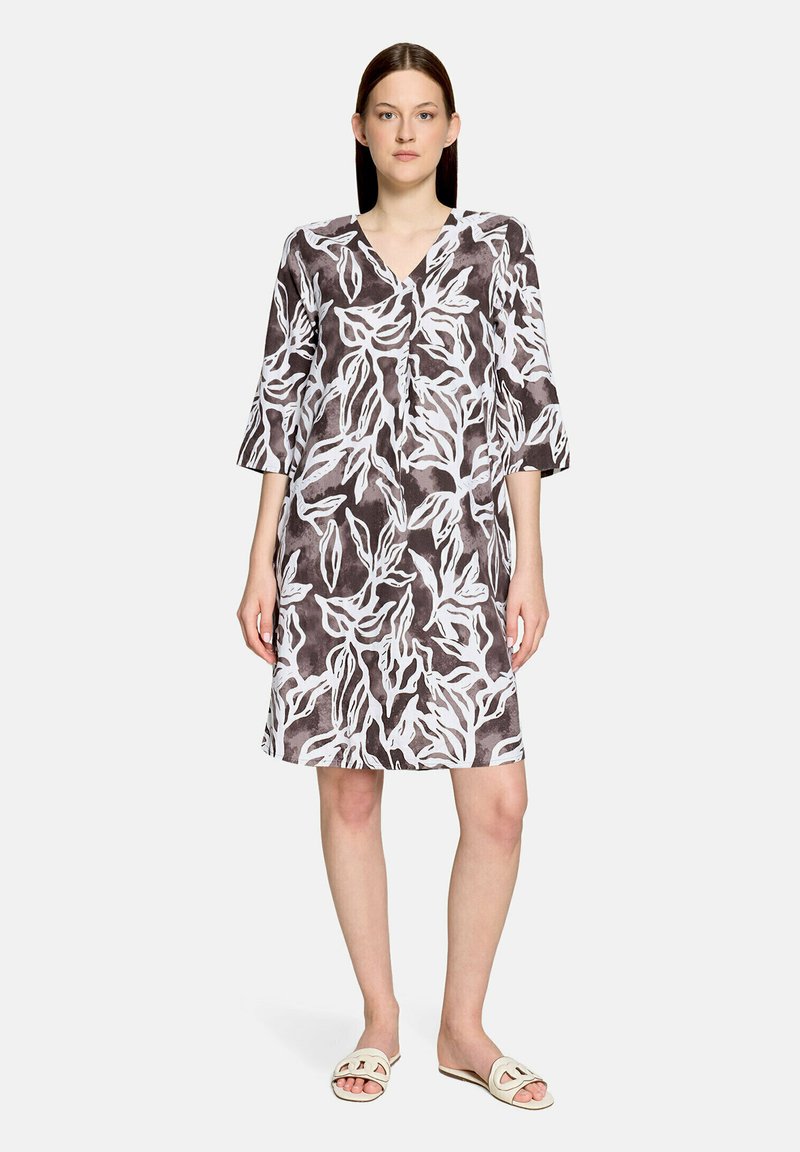 Femme debout portant une robe mi-longue à col en V avec des manches trois-quarts et un motif abstrait de feuilles blanches sur fond sombre, assortie à des sandales blanches.