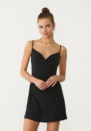 Robe noire avec un corsage ajusté, décolleté en V et bretelles spaghetti réglables. La jupe est légèrement évasée. Modèle portant des bijoux.