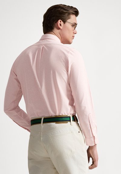 Ροζ πουκαμίσα με ρίγες, κλασικό γιακά και μακριά μανίκια, σε συνδυασμό με ανοιχτό μπεζ παντελόνι και πολύχρωμη ριγωτή ζώνη.