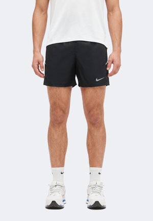 Muž oblečený v čiernych Nike šortkách, bielych Nike ponožkách a bielych bežeckých topánkach, stojaci pred jednoduchým svetlým pozadím.
