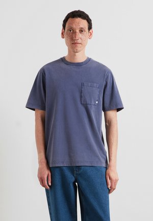 Giovane con capelli ricci e lentiggini che indossa una maglietta blu sbiadita a maniche corte con taschino sul petto e jeans blu, in piedi davanti a uno sfondo uniforme.