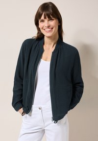Dunkelblau-grüne Bomberjacke aus strukturiertem Stoff mit Reißverschluss, Seitentaschen und gerippten Bündchen, kombiniert mit einem weißen Hemd und hellen Hosen.