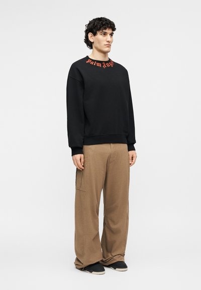 Νέος άνδρας με σγουρά μαλλιά που φοράει μαύρο φούτερ με κόκκινο κείμενο στη λαιμόκοψη, χαλαρά καφέ παντελόνια και μαύρα αθλητικά παπούτσια, στέκεται μπροστά σε λευκό φόντο.