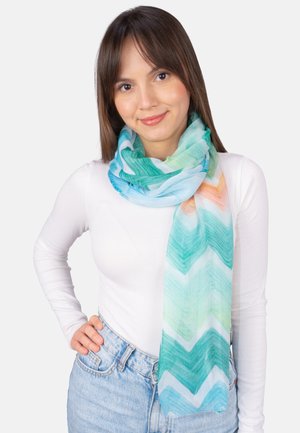 Junge Frau mit glatten braunen Haaren trägt ein weißes Langarmshirt, blaue Jeans und einen mehrfarbigen Schal mit Zickzackmuster.