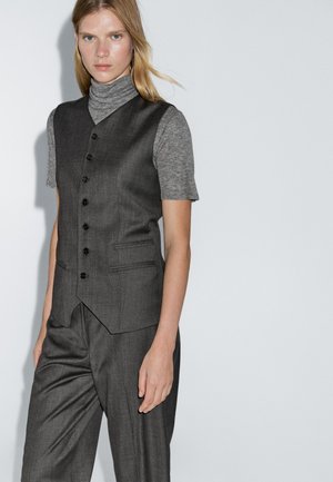 Gilet grigio su misura con colletto a punte, sei bottoni neri e tasche laterali, abbinato a una maglietta grigia chiara a maniche corte. Tessuto dalla texture liscia.