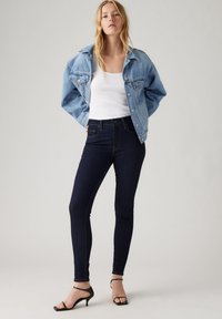 Kvinne iført lys blå denimjakke, hvit tanktop, mørk skinny jeans og svarte høye hæler som står mot en ensfarget bakgrunn.