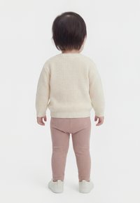 Småbarn set bagfra iført en cremefarvet striktrøje, lyserøde ribstrikkede leggings og hvide slip-on-sko på en hvid baggrund.