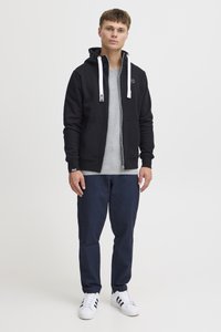 Sweat à capuche noir zippé avec cordons blancs, porté par-dessus un t-shirt gris. Assorti à un pantalon bleu marine et des baskets blanches avec des rayures noires.