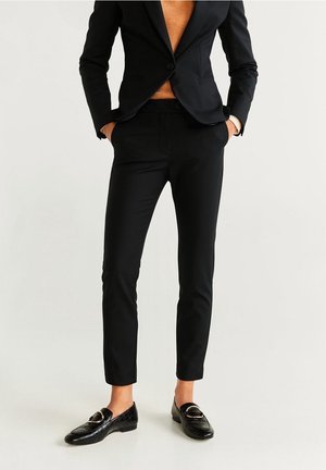 Frau trägt schwarze, maßgeschneiderte Hose, passendes Blazer, beigefarbenen Pullover und schwarze Loafers mit goldenen Schnallen, steht mit den Händen in den Taschen.