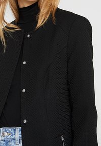 Chaqueta negra texturizada con botones de presión plateados y un bolsillo con cremallera, que presenta un diseño estructurado y un forro interior suave.