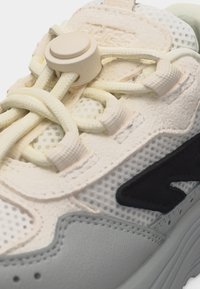 Közeli kép egy bézs színű tornacipőről, amely hálós anyagból készült, párnázott fűzőlyukakkal és lekerekített műanyag cipőfűzőzárral, amely vastag, krémszínű fűzőket tart rögzítve.