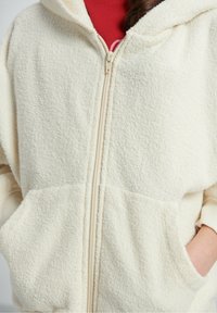 Sweat à capuche zippé en polaire de couleur crème avec tissu texturé, grandes poches avant et une capuche; doté d'une fermeture éclair crème et d'une doublure douce.