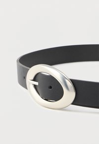 Svart lærbelte med en stor, glatt, oval formet sølvspenne som er festet gjennom ett hull på en enkel lys bakgrunn.