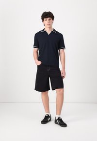 Mornarska polo majica s bijelim akcentima, uparen s crnim trapericama kratkih nogavica. Model nosi crne tenisice s bijelim trakama. Jednostavan, ležeran dizajn.