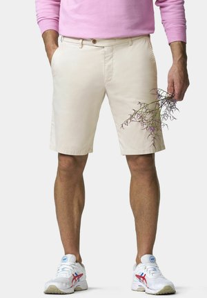 Mann trägt beige Shorts, weiße Sneaker mit roten und blauen Akzenten und ein pinkfarbenes Langarmshirt, hält einen kleinen Zweig mit violetten Blüten.