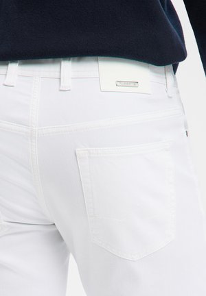 Gros plan sur la poche arrière et les passants de ceinture d'un jean en denim blanc, porté avec un haut foncé, montrant l'étiquette de la marque Alberto sur la ceinture.