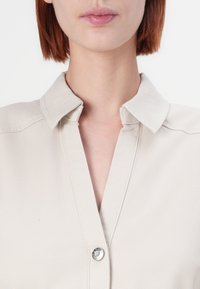 Chemise beige à col avec une fermeture à bouton, présentant un design épuré et une texture semblable au coton. Vue en gros plan montrant les détails de l'encolure et du col.