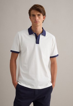 Giovane uomo in piedi con le mani in tasca che indossa una polo bianca con colletto e bordi delle maniche blu navy e pantaloni scuri su sfondo uniforme.