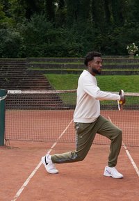 Sportininkas, apsirengęs balta ilgomis rankovėmis marškiniais ir alyvuogių žalumo kelniuose su juostelėmis, laiko teniso raketę ant molio aikštelės. Matomos baltos sportinės batelės.