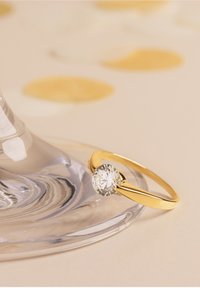 Anello d'oro con una pietra preziosa rotonda e trasparente incastonata in una fascia liscia, posato su una superficie di vetro con delicati accenti circolari sfocati sullo sfondo.