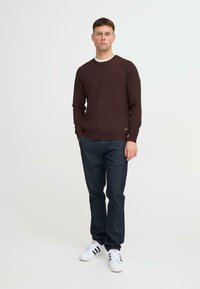 Bordeaux strikkegenser med V-hals, lange ermer, sammen med mørk denimjeans og hvite sneakers med svarte striper. Enfarget bakgrunn.