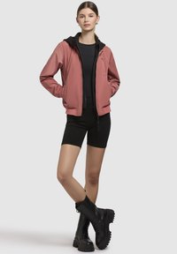 Veste bomber rose avec une capuche noire, fermeture éclair sur le devant et poignets côtelés, associée à un short noir et des bottes noires à semelles épaisses. Design simple et décontracté.