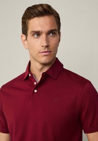 Burgundské polo triko s límečkem, krátkými rukávy a třemi knoflíky; obsahuje malé, nenápadné logo na hrudi. Hladká textura.