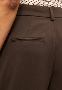 Κλείσιμο ενός ατόμου που φοράει σκούρο καφέ παντελόνια με τσέπη με ραφή στο πίσω μέρος και πουκάμισο με σχέδιο περασμένο μέσα.