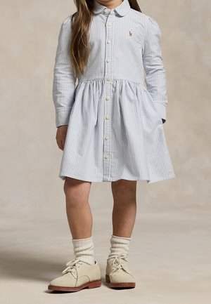 Kind draagt een lichtblauwe, lange mouwen, geknoopte jurk met zakken, crèmekleurige sokken en beige veterschoenen, staande op een neutrale achtergrond.