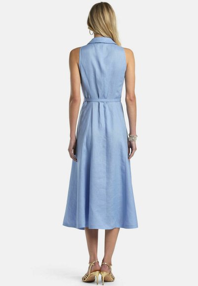 Žena v svetlomodrých midi šatách bez rukávov s golierom a priliehavým pásom, v páre so zlatými sandálmi na vysokom opätku, zobrazená zozadu.