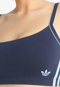 Marinblå sportbehå tillverkad av mjukt tyg, med ljusblå detaljer, en stilren design och en liten vit logotyp längst ner till vänster.