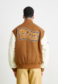 Smeđa i krema varsity jakna s rebrastim manžetama, "BROWN DUCKS" izvezeno velikim slovima na leđima. Glatka tekstura, opušten fit.