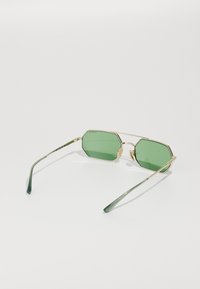 Ochelari de soare octagonali verzi cu un cadru din metal aurit, având vârful brațelor transparent verde și un design subțire, minimalist.