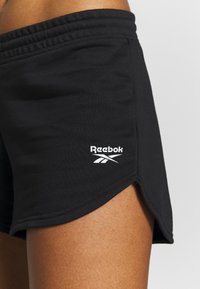 Bermudas de algodón negras con un dobladillo curvado, que presentan un logo de Reebok blanco en la parte inferior izquierda. La cinturilla es elástica para mayor comodidad.
