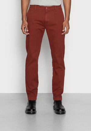 Uomo che indossa pantaloni dritti color ruggine e stivaletti in pelle nera, in piedi contro una parete bianca liscia.