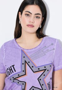 Camiseta de algodón púrpura con cuello redondo. Presenta un diseño gráfico de estrella con lunares negros y texto, sobre un fondo texturizado.