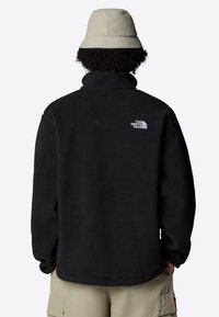 Svart fleecejacka med hög krage, med en liten vit logotyp på övre ryggen. Strukturerad tyg och avslappnad passform. Avslappnad klädsel.