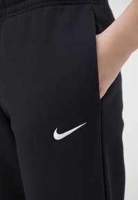 Pantalones de chándal negros hechos de tela suave, con un logo blanco de Nike en el lado izquierdo y bolsillos laterales para mayor funcionalidad.
