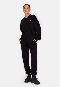 Melnā sporta krekls un bikses, izgatavotas no mīkstas auduma, ar mazu baltu logotipu uz krūtīm, brīva piegriezuma un ar šnoru jostu.