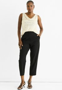 Woman wearing a sleeveless sequin beige top, black high-waisted cropped pants, and black strappy heels standing against a white background.