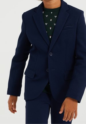 Enfant portant un costume texturé bleu marine avec un pull festif vert foncé orné de petits visages de Père Noël et de rennes.