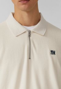 Hellbeige Reißverschluss-Polohemd mit geripptem Kragen und strukturiertem Stoff, das ein kleines navyfarbenes logopatch auf der linken Brust aufweist.