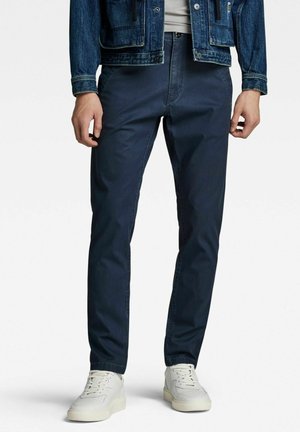 Hombre con pantalones ajustados azul oscuro, zapatillas blancas, camisa de color claro y chaqueta vaquera azul frente a un fondo liso.